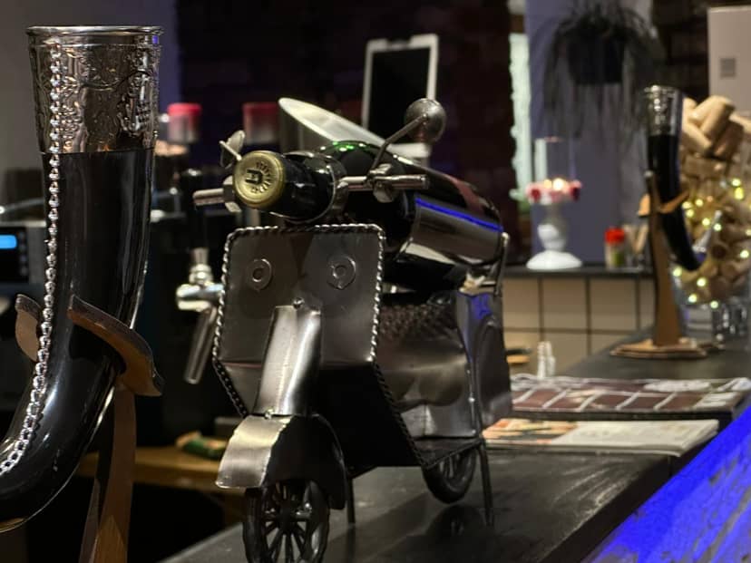 Ein Detailfoto zeigt dekorative Objekte auf einer Bar, darunter ein Weinflaschenhalter in Form eines Metallrollers und ein verzierter Trinkhorn.