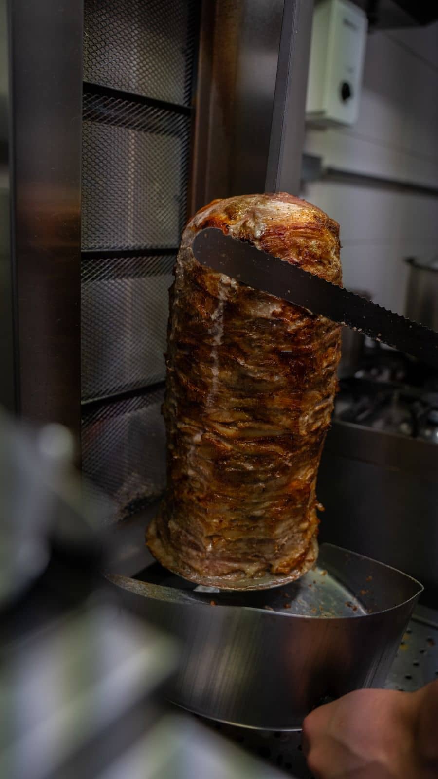 Ein großer Fleischspieß (Döner/Gyros) wird mit einem gezackten Messer geschnitten, während eine Hand darunter eine Auffangschale hält.