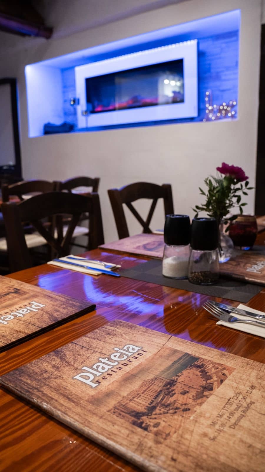 Ein gedeckter Restauranttisch mit Menükarten, Besteck und Gewürzstreuern steht im Vordergrund, während im Hintergrund eine Nische mit blauer LED-Beleuchtung…