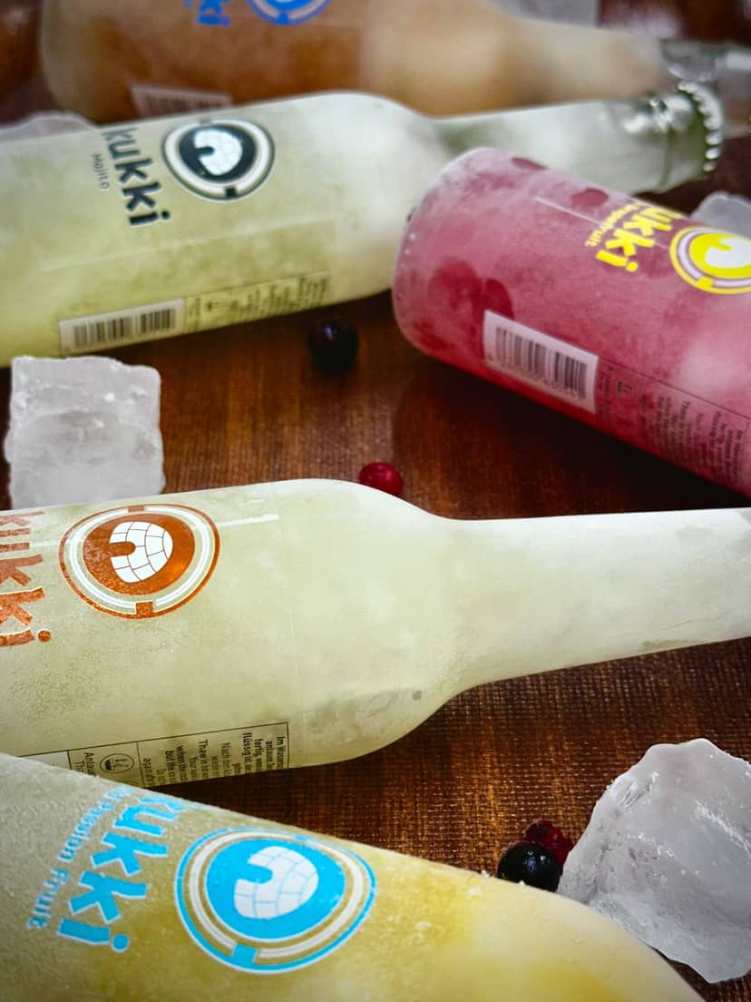 Das Foto zeigt mehrere gefrorene Kukki-Flaschen mit verschiedenen Geschmacksrichtungen wie Mojito und Passion Fruit, umgeben von Eiswürfeln und Beeren auf…